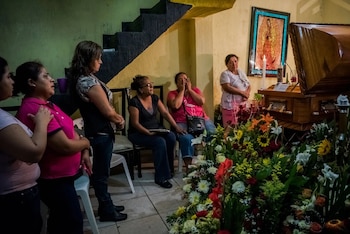 Seis mujeres en un velatorio junto a un ataúd de madera, arreglos florales, una imagen religiosa, una vela encendida y una escalera
