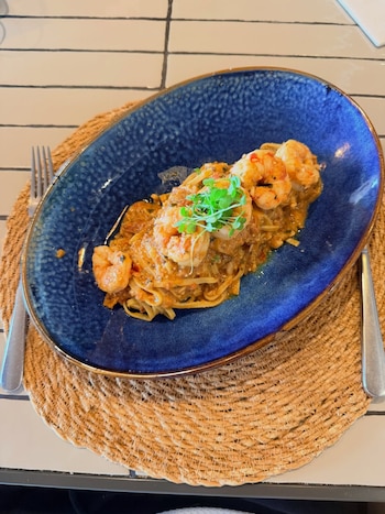 Plato ovalado azul con pasta, camarones cocidos, salsa de tomate y brotes verdes. Un tenedor de metal a la izquierda. Un mantel individual tejido de rafia debajo del plato