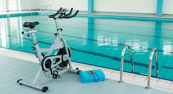 Bicicleta fija de ejercicio sobre el suelo junto a una piscina cubierta, con una toalla azul y un monitor de ritmo cardíaco apoyados en el piso.