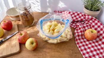 Una bolsa con manzanas troceadas, manzanas enteras, un cuchillo sobre una tabla, un frasco con canela y un paño a cuadros en una mesa de madera.