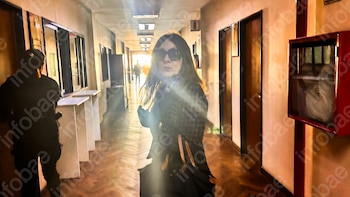 Pasillo largo, mujer de pelo oscuro con gafas de sol en primer plano, persona uniformada de espalda, puertas, ventanas, y caja roja en pared