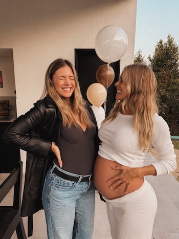Dos mujeres rubias, una sonriendo con ojos cerrados y la otra embarazada tocándose el vientre, posan junto a globos de colores claros en el exterior