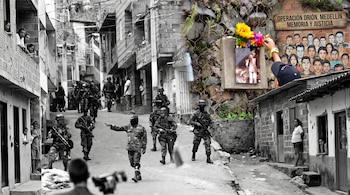 Soldados armados patrullan una calle empinada mientras vecinos observan; a la derecha hay un mural con rostros y un altar con flores.