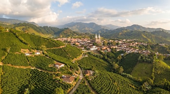 Vista aérea de un pueblo rodeado por colinas verdes con plantaciones de café en terrazas, caminos de tierra, casas dispersas y una iglesia central.