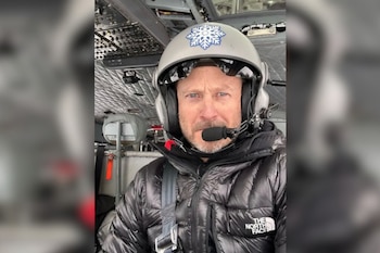 Un hombre con casco gris, micrófono y chaqueta negra mira a la cámara desde el interior de la cabina de un helicóptero