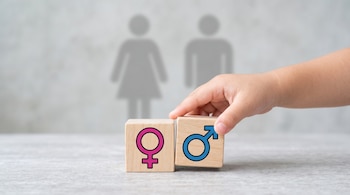 Una mano infantil junta dos cubos de madera con los símbolos femenino y masculino frente a siluetas borrosas de una mujer y un hombre.