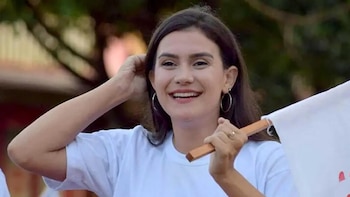 Gabriela De Paiva, mujer sonriente con cabello oscuro y largo, viste camiseta blanca y sostiene una pequeña bandera blanca. Fondo verde borroso