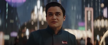 Primer plano de Katy O’Brian con cabello oscuro y corto, vistiendo una chaqueta azul marino con cuello alto y una insignia. El fondo borroso muestra luces de ciudad nocturna