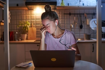 Joven mujer con cabello recogido, auriculares, y gafas, frente a una laptop en una cocina con azulejos, encimera, utensilios y plantas