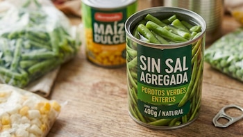 Una lata de porotos verdes con etiqueta "Sin Sal Agregada", una lata de maíz dulce, bolsas de porotos verdes, arvejas y maíz congelados, y anilla de lata sobre madera