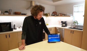 Un hombre de cabello rizado con camiseta negra está de pie en una cocina frente a una tablet sobre una encimera. Se observan electrodomésticos