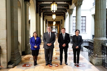 Cuatro personas vestidas con trajes formales están de pie en un pasillo con columnas de piedra, piso de mosaico y lámparas colgantes