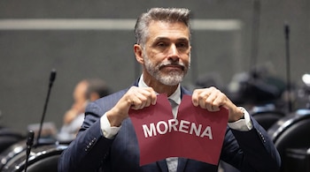 Un hombre de traje, con cabello y barba grises, mira al frente mientras rompe con ambas manos una hoja de papel color guinda con la palabra 'MORENA'.