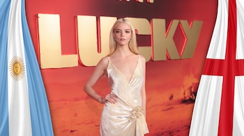 Anya Taylor-Joy con cabello rubio y vestido color crema, posa frente a un letrero con la palabra LUCKY, entre las banderas de Argentina e Inglaterra.