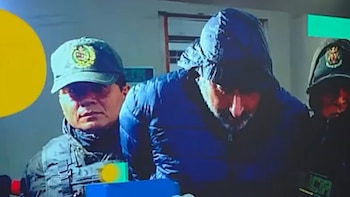 Un hombre con chaqueta azul y capucha agacha la cabeza mientras dos policías con gorra militar lo sujetan por los brazos