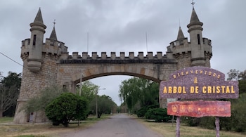 Arcada de piedra con dos torreones y almenas sobre un camino. Un cartel a la derecha indica "Bienvenidos al Árbol de Cristal". Vegetación verde a los lados.