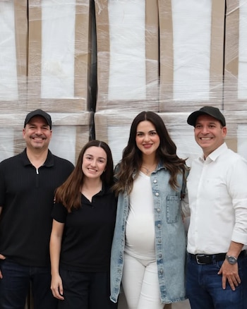 Cuatro personas, incluyendo a Andrea Chávez, sonríen frente a pilas de cajas embaladas; la mujer central embarazada viste chaqueta de mezclilla
