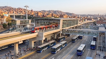 Una estación de tren elevado con un tren rojo y blanco, una terminal de autobuses en el nivel inferior y un teleférico sobre una colina
