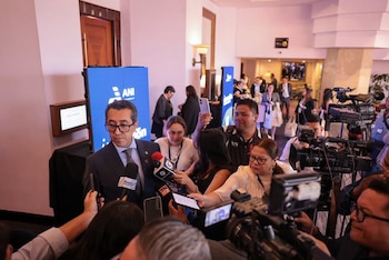 Un hombre con traje oscuro y gafas habla a varios micrófonos. Está rodeado de periodistas, cámaras de video, teléfonos y una pantalla azul con el logo ANI