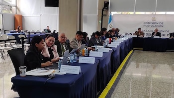 Sala de conferencias. Mesa larga cubierta con personas sentadas, botellas de agua, papeles, micrófonos y etiquetas de nombre. Banner y bandera de Guatemala