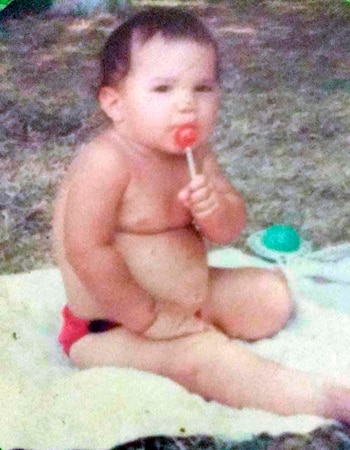 Un bebé de pelo oscuro y pañal rojo, sentado en una toalla clara, chupando una piruleta roja con la mano derecha, con fondo borroso de hierba