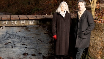 Una mujer de cabello blanco y un hombre con bufanda clara, ambos con abrigos oscuros, posan junto a un arroyo con un puente de madera al fondo