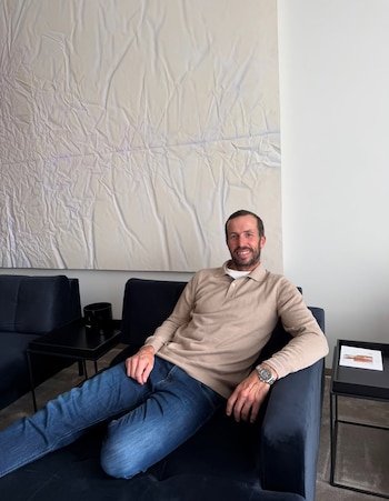 Un hombre con suéter beige y pantalón azul permanece sentado en un sillón oscuro frente a un gran Cuadro blanco en una pared