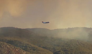 Una pequeña aeronave vuela sobre colinas cubiertas de vegetación y un denso humo en un cielo nublado