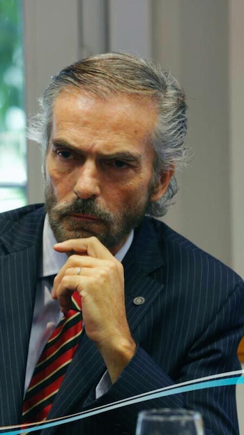 Primer plano del juez Gustavo Hornos, un hombre con cabello canoso y barba, vistiendo un traje a rayas y corbata roja, con la mano en la barbilla