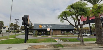 Una vista exterior de un restaurante Carl's Jr gris con una estrella amarilla, un letrero de drive-thru, árboles, palmeras y una calle con vehículos bajo un cielo nublado