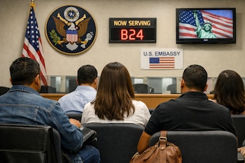 Personas sentadas de espaldas en una sala de espera con símbolos de EE. UU. en la pared: una bandera, el Gran Sello, un letrero digital y una señal de 'U.S. Embassy'.
