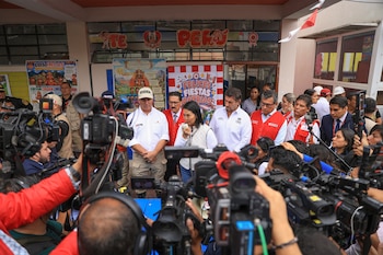 Una mujer habla por micrófono rodeada de varios hombres y cámaras de video. Al fondo se ven ventanas con carteles educativos sobre Perú y Fiestas Patrias