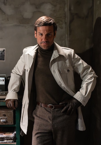Hombre de mediana edad con cabello oscuro y patillas, vestido con gabardina clara, jersey de cuello alto marrón y guantes, con expresión seria