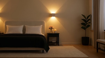 Cama con cabecero tapizado, dos almohadas blancas, edredón negro, mesita de noche con florero de rosas, vela encendida, lámpara de pared, planta en maceta y ventana.
