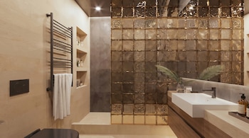 Un baño moderno con una pared decorativa de bloques de vidrio dorados, un lavabo blanco sobre un tocador de madera oscura y un toallero negro con una toalla blanca.