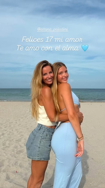 Dos mujeres sonríen abrazadas en una playa con el mar y el cielo azul de fondo. Una mujer usa top blanco y shorts de jean; la otra, bikini y pantalón celestes