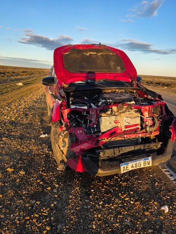 Automóvil rojo con la parte delantera destruida y el capó abierto al costado de una ruta asfaltada en una zona rural