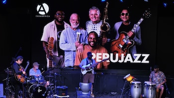Cinco músicos tocan en un escenario; detrás, una pantalla proyecta una foto de cinco hombres y los textos 'CASAMÉRICA' y 'PERUJAZZ'