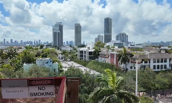 Vista aérea de un paisaje urbano en el sur de Florida, mostrando rascacielos, edificios residenciales, densa vegetación y palmeras. Un cartel de 'PELIGRO NO FUMAR' en primer plano