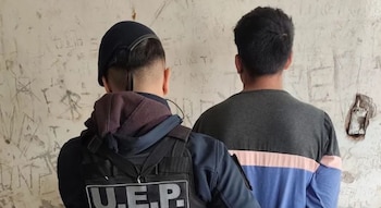 Un agente policial y un hombre de espaldas están de pie frente a una pared clara con múltiples marcas e inscripciones