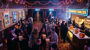 Personas con disfraces, calabazas iluminadas, carteles de cine y un mostrador de palomitas en el vestíbulo de un cine con alfombra estampada