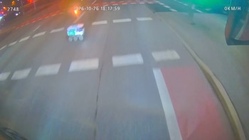 Calzada nocturna con marcas viales discontinuas por donde avanza un pequeño robot de reparto con ruedas frente a un vehículo