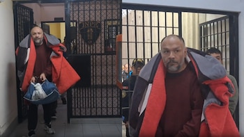Un hombre con barba, camiseta vinotinto, manta gris y roja, y manos esposadas sostiene una bolsa azul, ingresando a un edificio con rejas y el logo del INPE