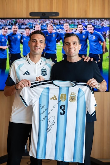 Dos hombres sostienen una camiseta de fútbol con rayas celestes y blancas, el número nueve, el escudo de AFA y el logo de FIFA, frente a una pantalla con personas