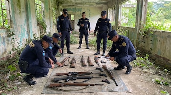 Cinco policías de Guatemala inspeccionan once armas de fuego: fusiles, escopetas y pistolas, sobre una lona en un edificio abandonado.