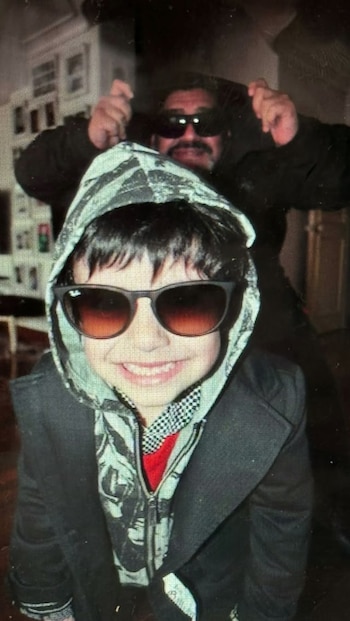 Un niño con capucha y gafas de sol sonríe en primer plano mientras un hombre con barba y gafas oscuras posa detrás de él en un interior