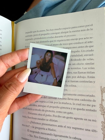 Una mano sostiene una fotografía instantánea que muestra a Antonela Roccuzzo sonriendo, con un mate y un libro. De fondo, un libro abierto y un marcapáginas