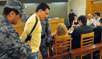 Hombre en camisa amarilla escoltado por oficiales camuflados en un tribunal. Se observan otras personas sentadas y personal de seguridad al fondo