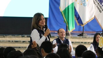 Una mujer habla en micrófono ante una audiencia. Un hombre sonríe sentado detrás. Al fondo hay tres banderas y una pantalla. Hay gente con manos levantadas