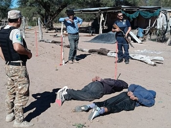 Dos hombres esposados boca abajo en el suelo de arena, custodiados por dos oficiales armados, con un civil y estructuras rurales al fondo